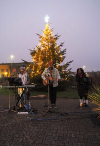A lit-up Christmas tree with three people playing a guitar, a keyboard, and singing in front of it in the evening