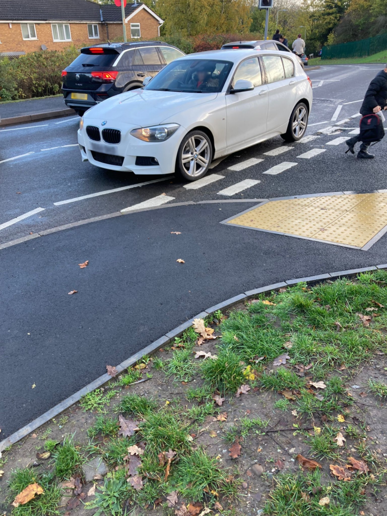 A photograph of a white car that has stopped across the width of a junction, blocking access to other vehicles