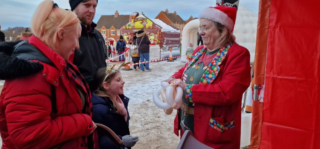A woman in a Santa hat twists balloons into shapes for an onlooking child
