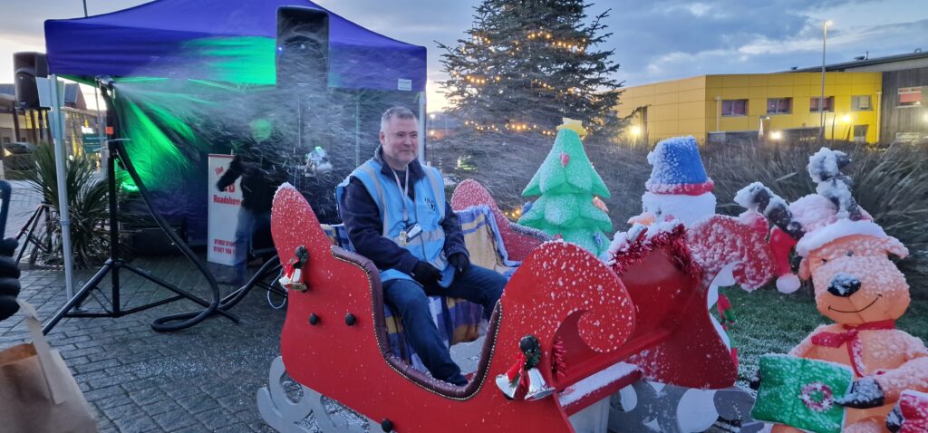 A man in a high-vis vest sits in Santa's sleigh, as artificial snow sprays all over him.