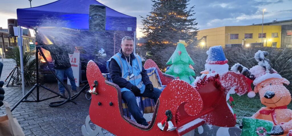 A man in a high-vis vest sits in Santa's sleigh, as artificial snow sprays all over him.