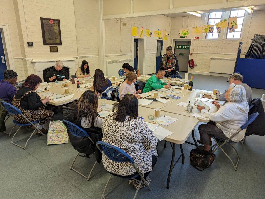 A group of people sitting around two large tables, all painting on sheets of paper