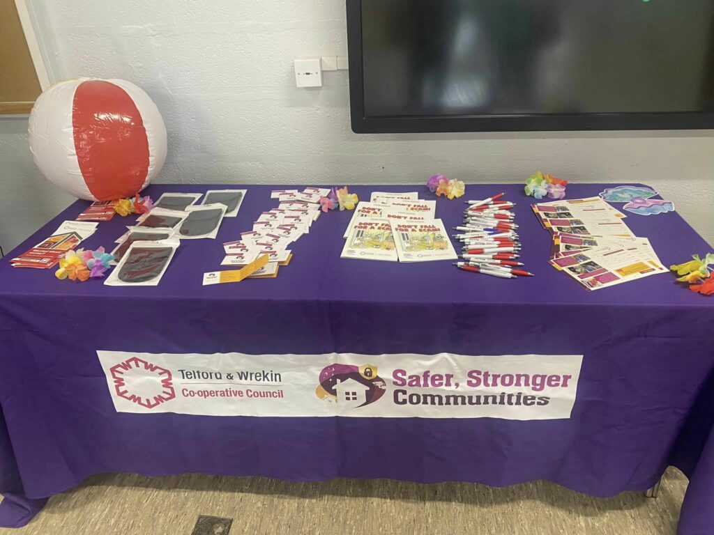 A photo of a table covered with a table cloth, a sign that reads "Safer Stronger Communities", and adorned with promotional pens and leaflets