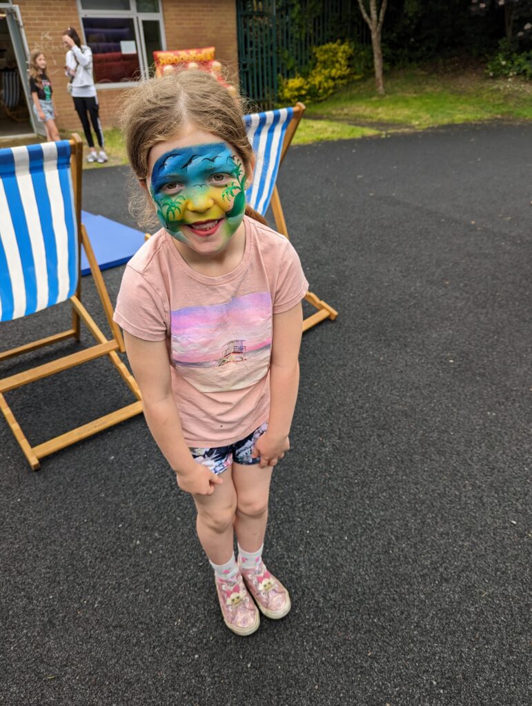 A child with full face paints standing in front of two deck chairs outside, smiling for the camera