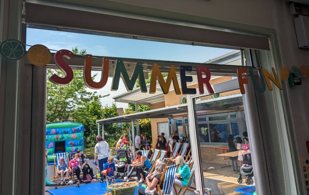 The words "summer fun" draped across the top of an open door. Behind are an assortment of deck chairs with people sitting outside, and an inflatable toy at the back