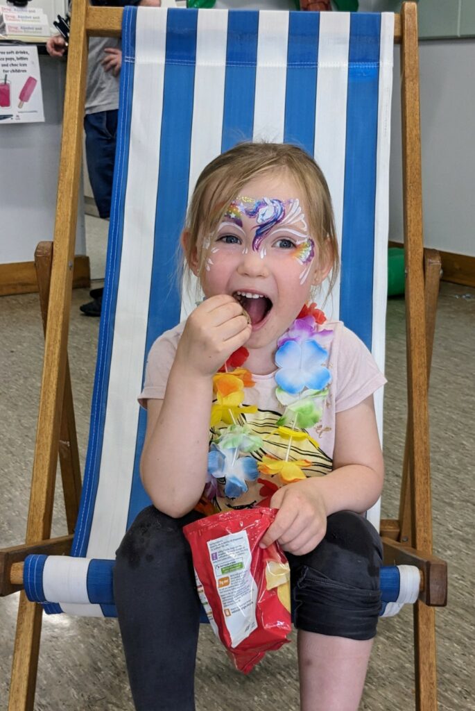 A child sitting in a deck chair eating crisps, with full face paints