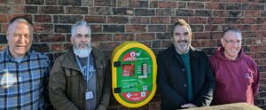 4 people standing beside the defibrillator unit, against a brick wall